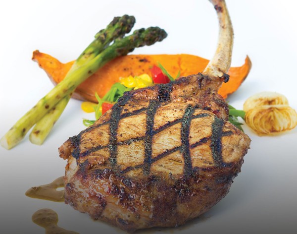 Caribbean-spiced, grilled Nagano pork chop with honey and cider vinegar sauce on a white plate with a side of sweet potato and asparagus spears.