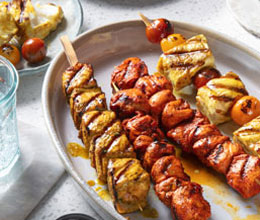 Three Indian-inspired kabobs including flaky white fish, chicken and blistered tomatoes with crushed chilis off to the side in a blue dish.