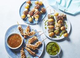 The South American-inspired kabobs, one with beef and potato, one potato and garlic, the last pork on blue plates with a bowl of chimichurri to the side. 