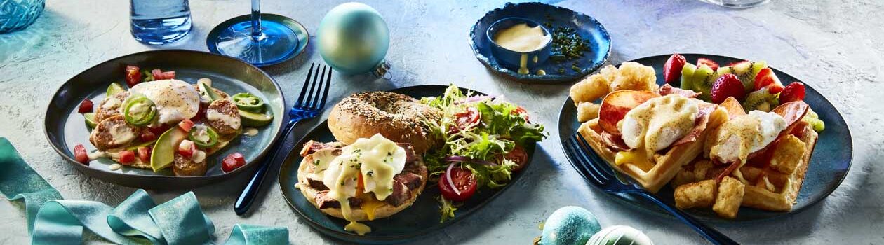 Three blue plates featuring three different kinds of eggs Benny recipes including one featuring waffles, another a NY-style bagel and one on a flour tortilla over a blue background.