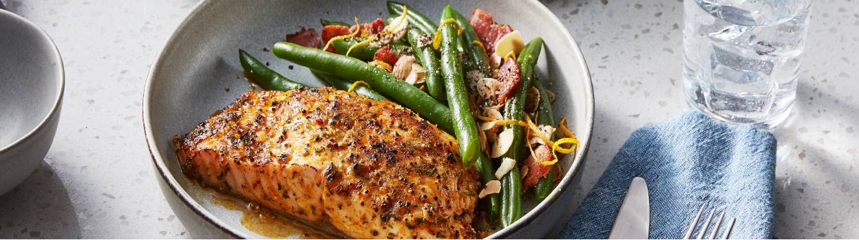 A marinated salmon fillet in a grey bowl next to a side of green beans flecked with almonds and lemon zest.