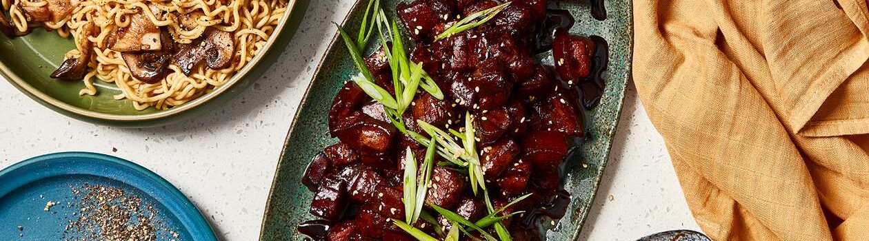 Mahogany lacquered pork belly cubes on a dark sage green oval serving platter next to a bowl of ramen noodles with mushrooms.