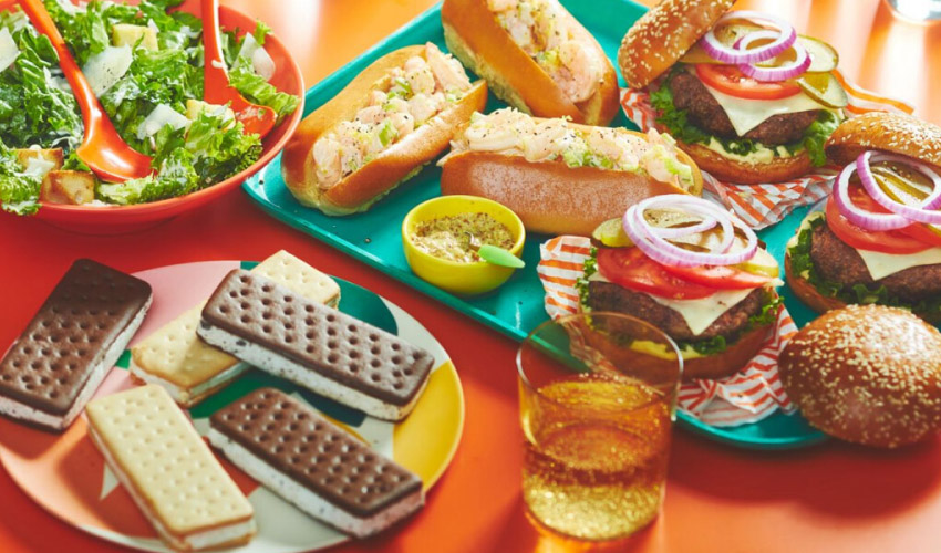 orange surface with platter of summer foods: shrimp rolls, ice cream sandwiches, caesar salad, burgers