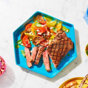 Sliced steak on an octagonal turquoise plate with a side of fresh tomato salad and a yellow plate with a Nagano pork chop and slaw on a white marble counter.