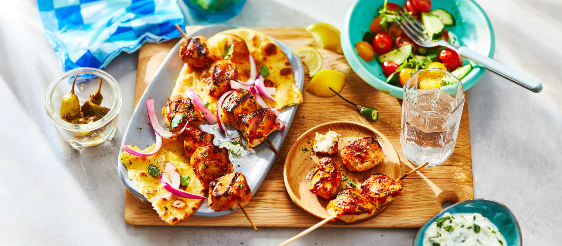 Grilled chicken skewer placed over a grilled naan flatbread on a blue platter sitting atop a wooden cutting board.