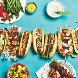 A row of variously dressed sausages and hot dogs, topped with roasted corn salad, various condiments, and fresh and pickled veggies, on a blue background with plates and bowls of fresh topping choices, including a mango salad.