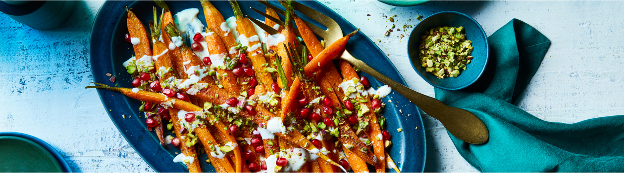 Oval blue platter of roasted carrots with yogurt tahini sauce, chopped pistachios, and fresh pomegranate arils