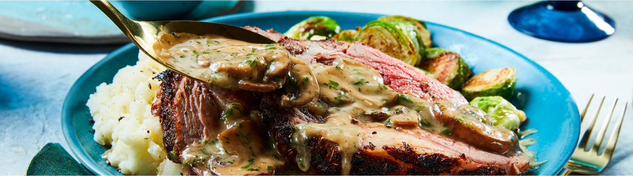 Round blue dinner plate with mashed potatoes, Brussels sprouts and Mushroom-Crust Prime Rib with Stroganoff Sauce being spooned overtop