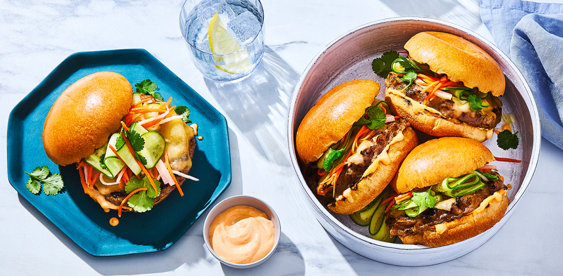 serving tray of three burgers, plus plated burger to the side, all stacked with a prime rib beef burger patty, pickled vegetables, cheese, and sriracha aioli on a brioche bun.