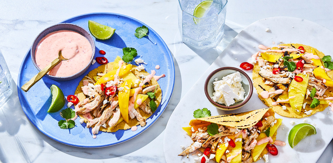 Serving board with tropical chicken and mango tacos, plus blue dinner plate to the side with chicken taco and bowl of chili-lime crema.