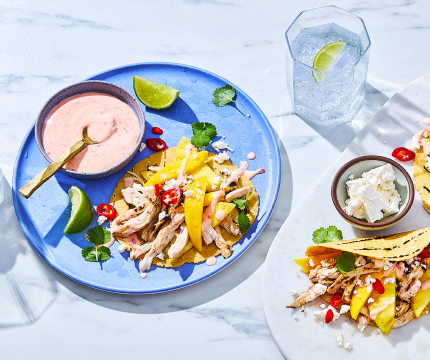 Serving board with tropical chicken and mango tacos, plus blue dinner plate to the side with chicken taco and bowl of chili-lime crema.