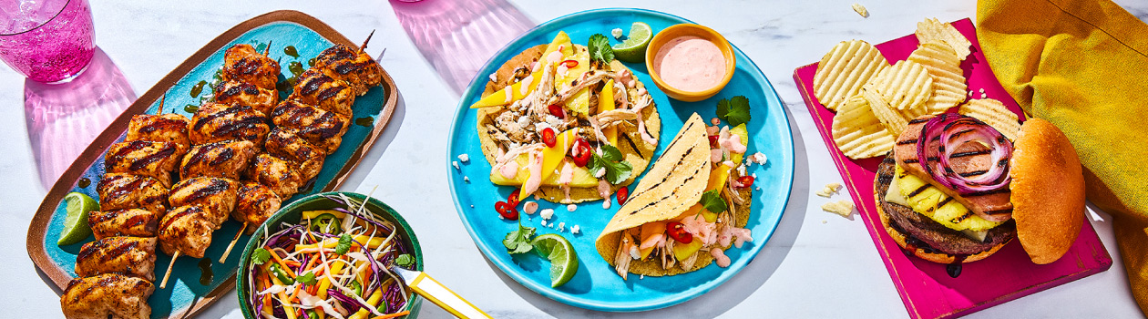 Marble surface with three platters of tropical-inspired food: Chicken kabobs, mango tacos and hawaiian-style burgers.